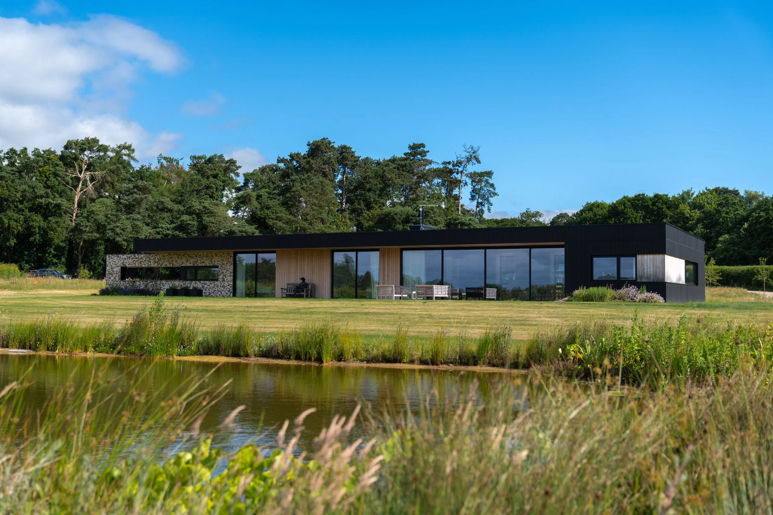 Modern house with large glass windows situated in a lush green area, overlooking a pond with tall grass in the foreground, surrounded by tall trees under a blue sky with clouds.