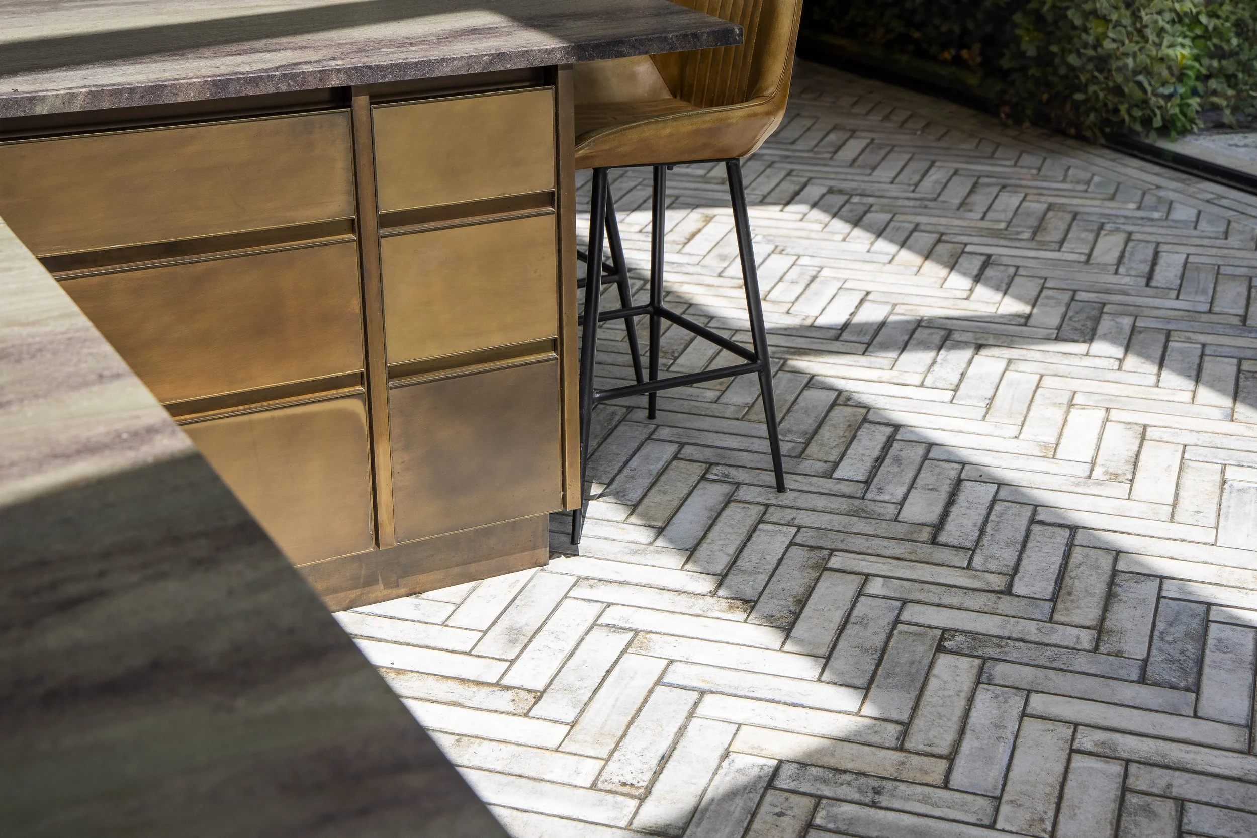 Close-up of a metallic kitchen island with a stone countertop, black metal barstool with wooden seat, and herringbone patterned stone patio beside lush green bushes.