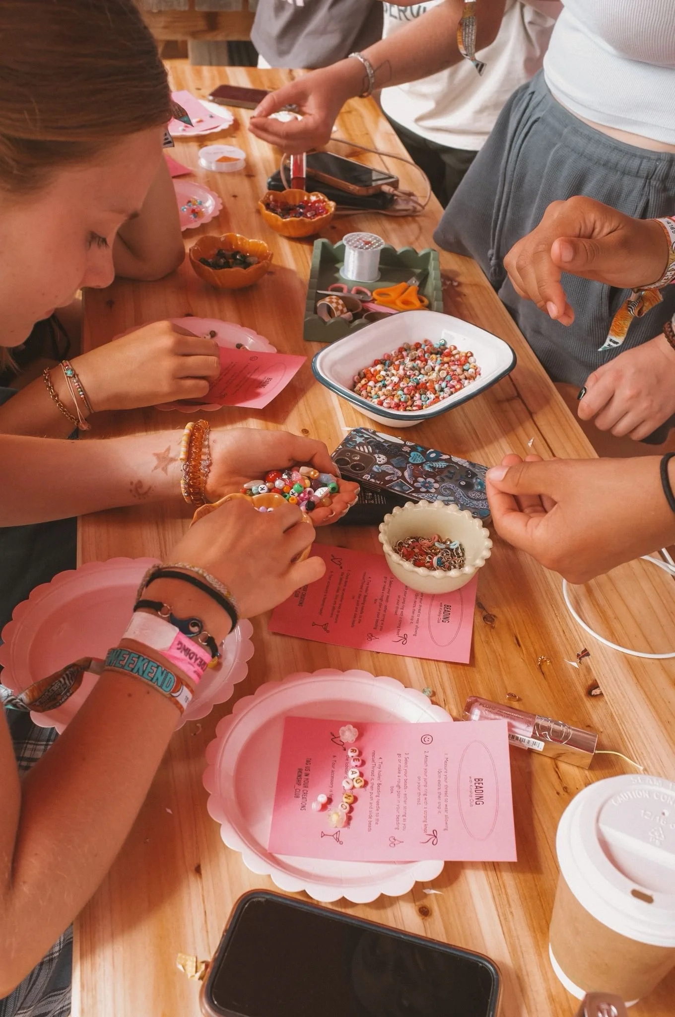 People participating in a bead craft activity at a wooden table, with pink paper placemats, bowls of beads, scissors, and crafting supplies scattered around.