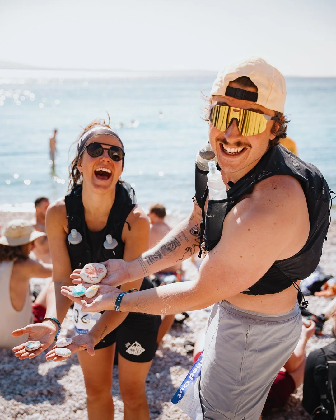 Smiling young man and woman on a beach holding decorated rocks, enjoying a sunny day with other beachgoers in the background.