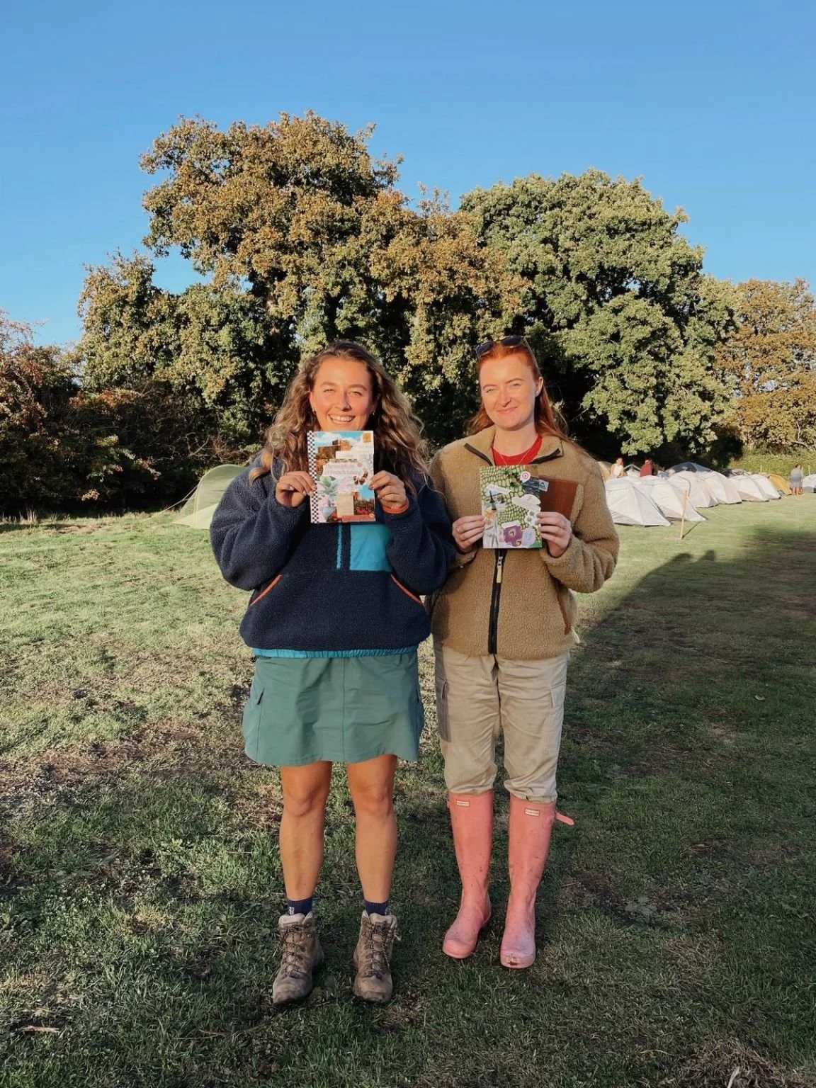 Two women standing outdoors in front of a large tree, holding colorful cards or brochures, with tents visible in the background on a grassy field during daytime.