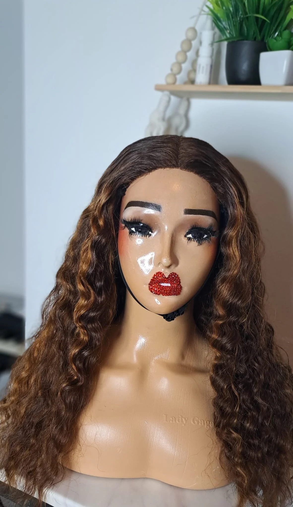 A mannequin head with long, curly brown hair, makeup including bold eyebrows, long eyelashes, and lips adorned with red rhinestones, set against a plain background with a shelf holding plants in black and white pots.