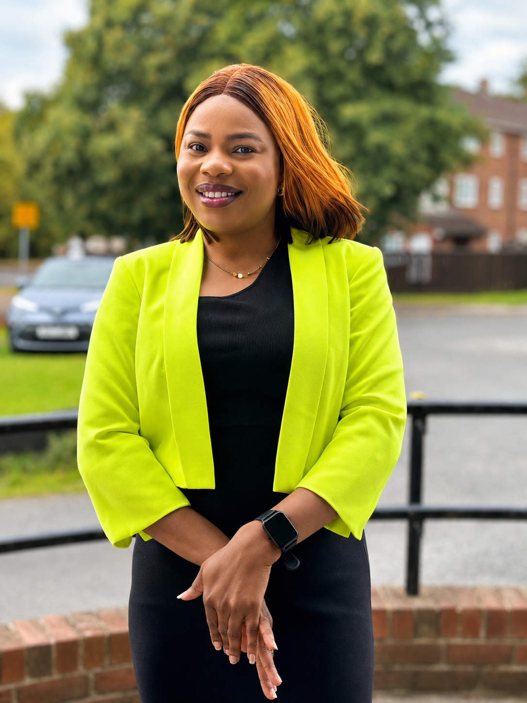 A confident woman with shoulder-length, reddish-brown hair, wearing a bright yellow blazer over a black top, standing outdoors in a residential area with cars, trees, and a brick building in the background.