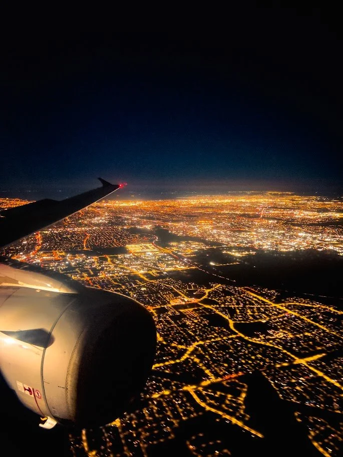 Nighttime aerial view of a city illuminated with bright lights, seen from an airplane window with part of the jet engine and wing visible in the foreground.