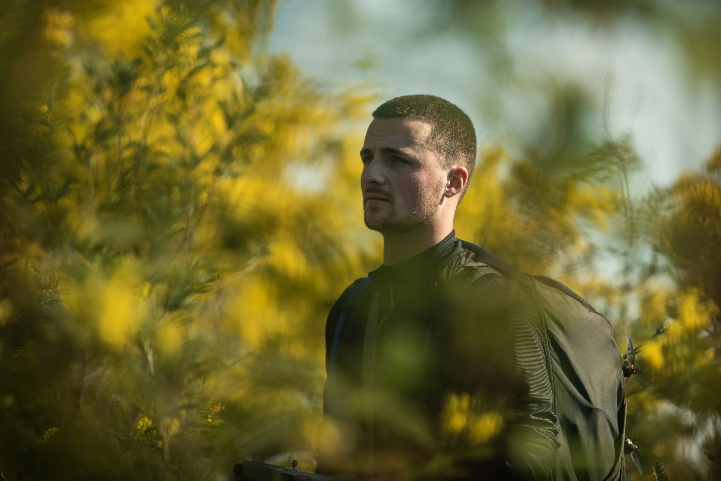 Un homme portant un sac à dos regarde vers l'avant dans un environnement naturel avec des feuillages verts et jaunes.
