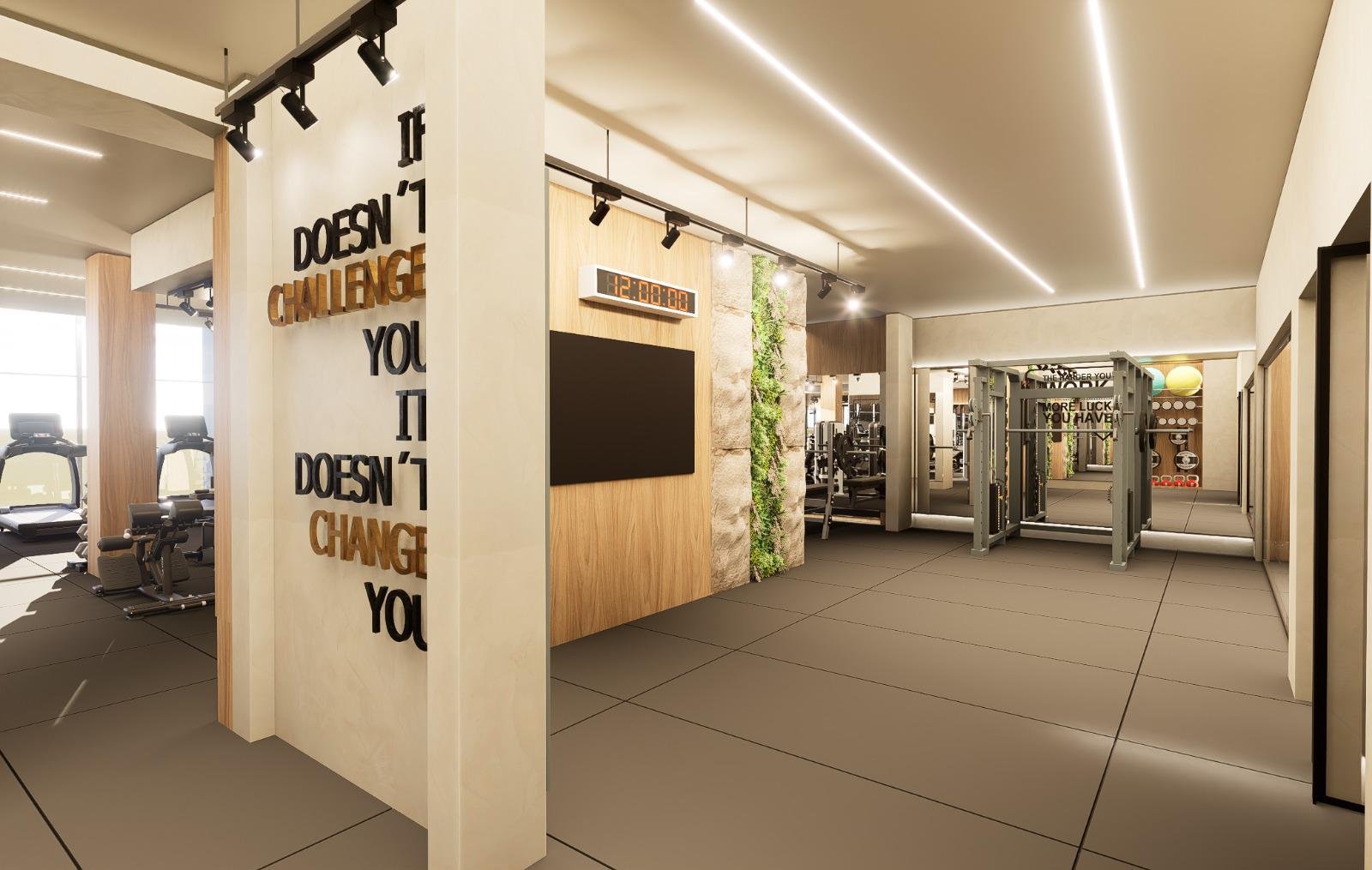Interior de academia moderna com equipamentos de treino, paredes decoradas, painel de TV, relógio digital e iluminação contemporânea.