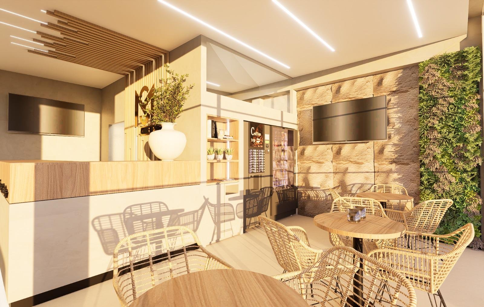 Interior de um ambiente de café com mesas de madeira, cadeiras de vime, paredes de pedra e uma parede com vegetação, iluminação moderna no teto, televsor na parede, balcão com máquina de bebidas e uma planta em um vaso branco.