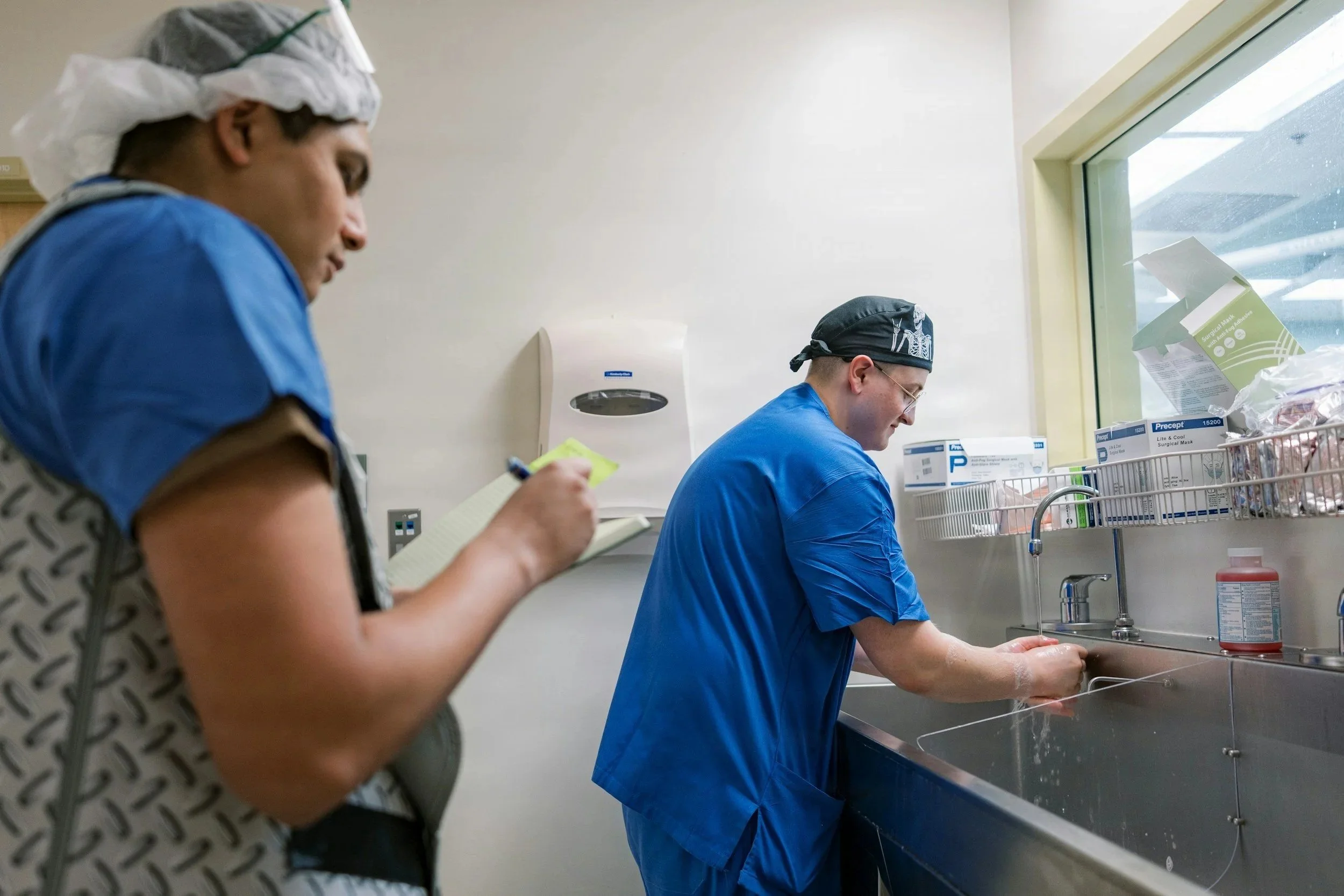 Two healthcare workers in scrubs and surgical caps washing their hands in a hospital sink.