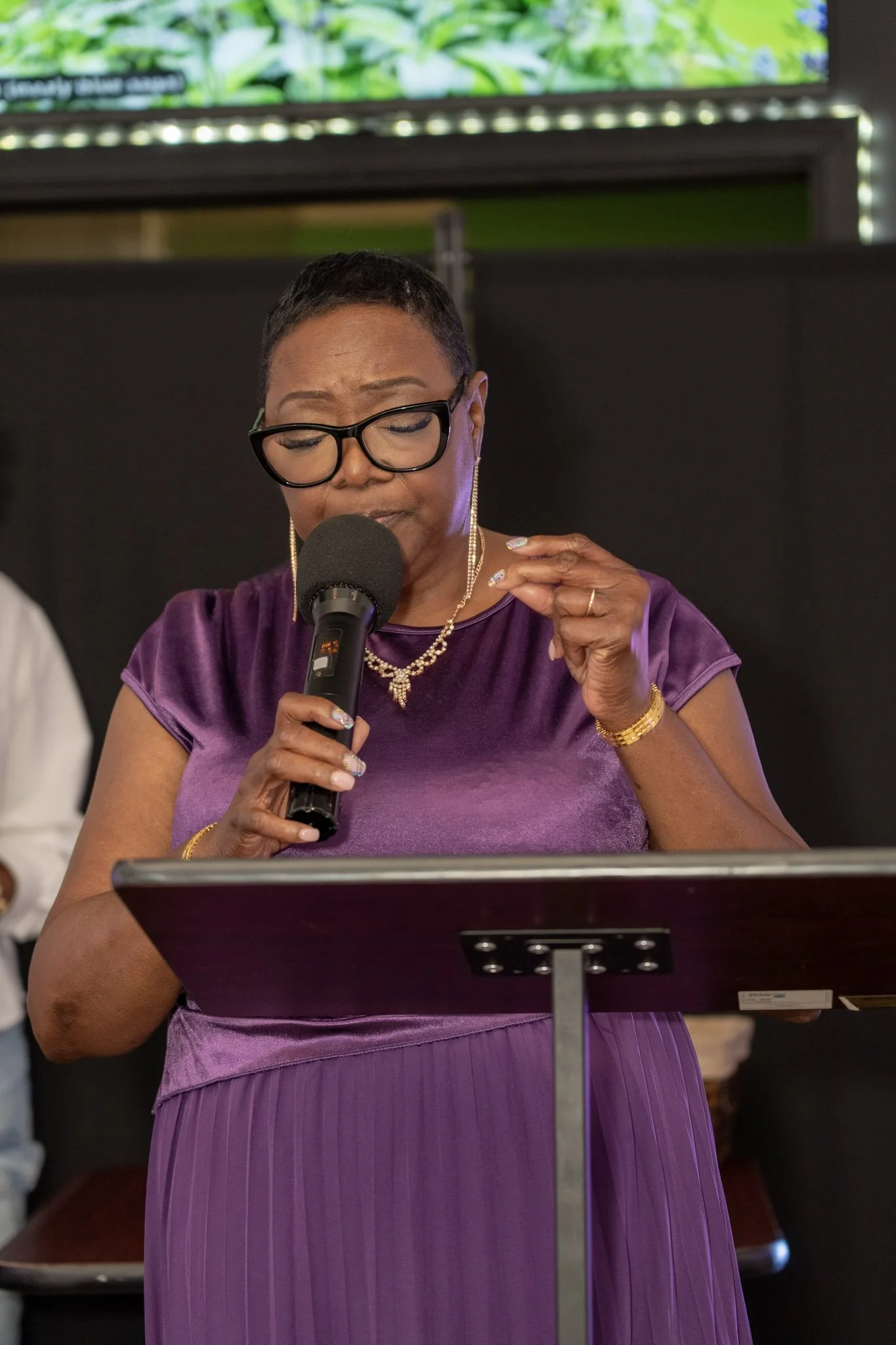 An African American woman with short hair, glasses, and gold jewelry, is speaking into a microphone while looking at a music stand or podium, wearing a purple dress, with trees and a screen in the background.