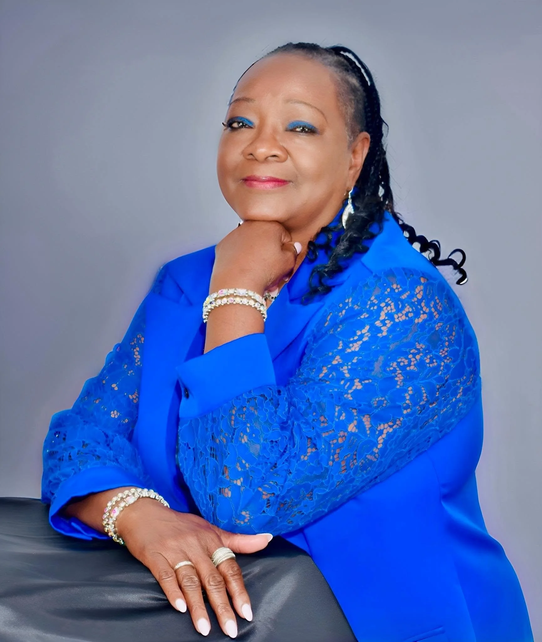 A woman wearing a bright blue lace blouse and matching jewelry, with a confident expression, resting her chin on her hand.