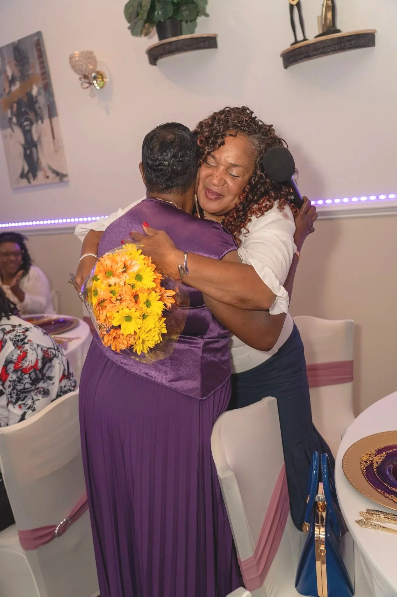 A woman with curly hair hugging a man during a celebration or event. The woman is holding a bouquet of yellow and orange flowers and has her eyes closed with a content expression. The setting includes a decorated table, chairs, and wall shelves with decorative items in the background.