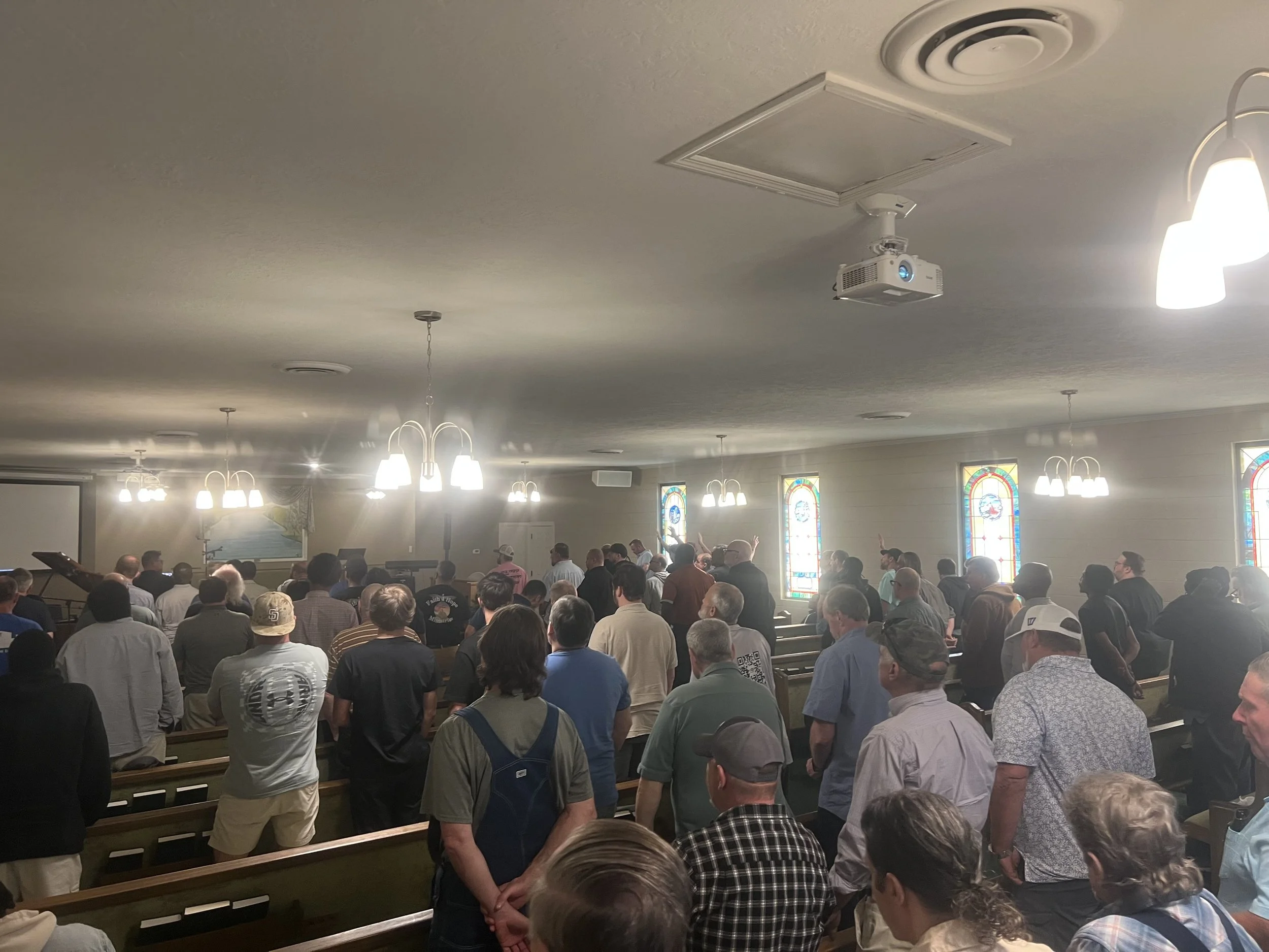 Mens meeting men worshipping the Lord, ministering to one another, revival. People gathered inside a church during a service or event, facing forward, with stained glass windows and ceiling lights visible.