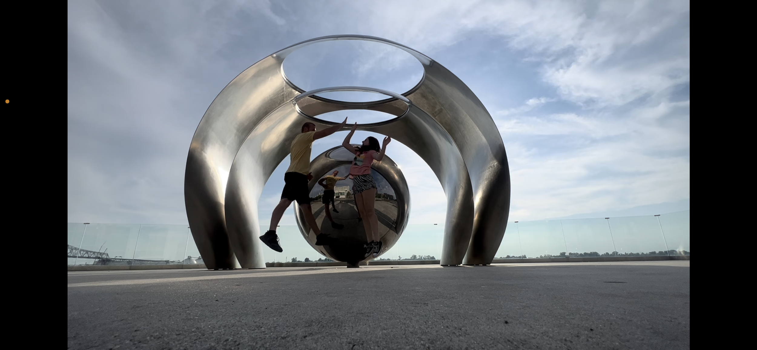 Two people, a man and a woman, high-fiving inside a large, shiny, metallic sculpture of interconnected rings and spheres, with a cloudy sky and distant landscape in the background.