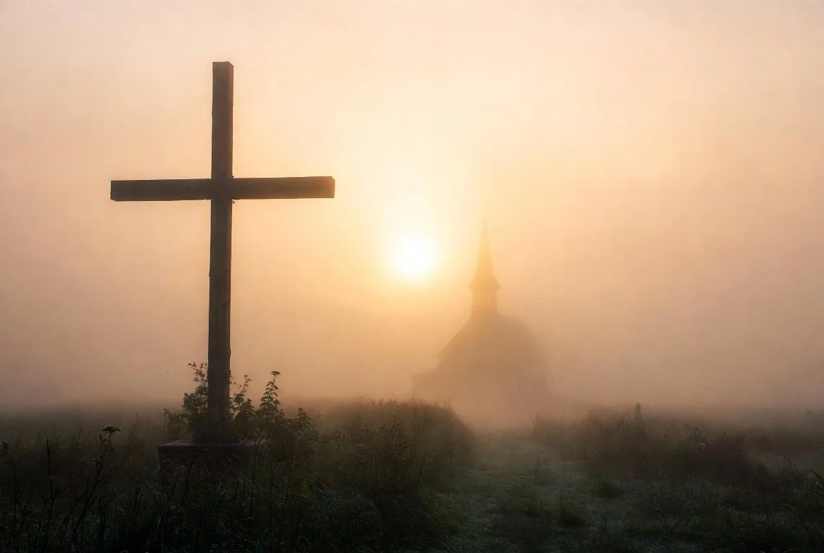 A cross in the foreground with a church in the background, through fog with the Sun shining behind.