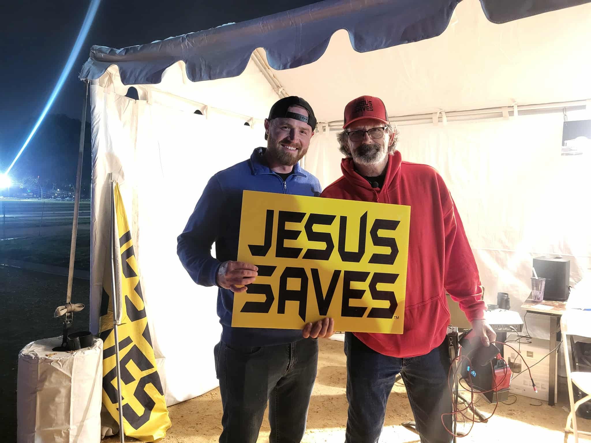 Two men one being Brother Caleb Shuford holding a yellow sign that says 'JESUS SAVES' stand inside a lit tent at night in Washington DC during Communion America for all 50 states revival celebrating America . 
