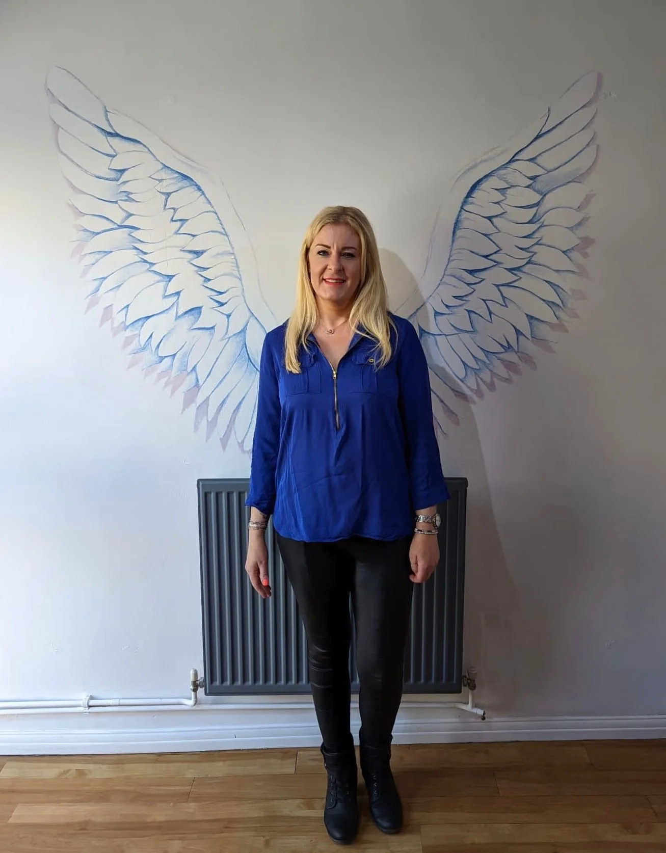 A woman with blonde hair wearing a blue zip-up jacket and black pants standing in front of a wall with large, artistic angel wings drawn on it.