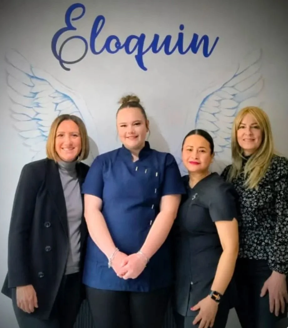 Four women standing in front of a wall with the word 'Eloquin' written in blue and angel wing illustrations. The women are smiling and dressed in professional attire, with one woman in the center wearing a blue medical uniform.