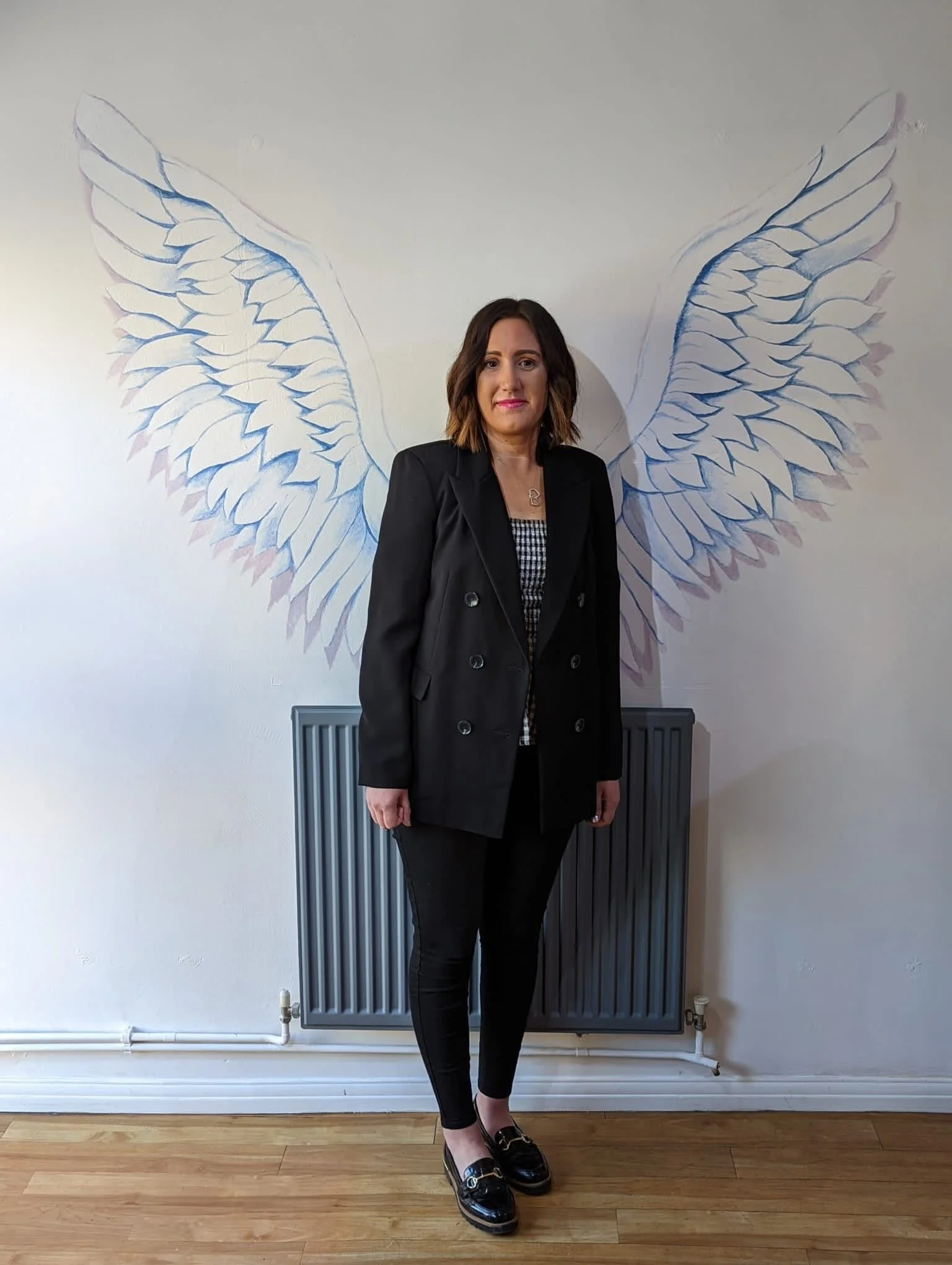 A woman with shoulder-length brown hair, dressed in a black blazer, checkered top, black pants, and black loafers, standing in front of a wall with mural of angel wings drawn behind her.