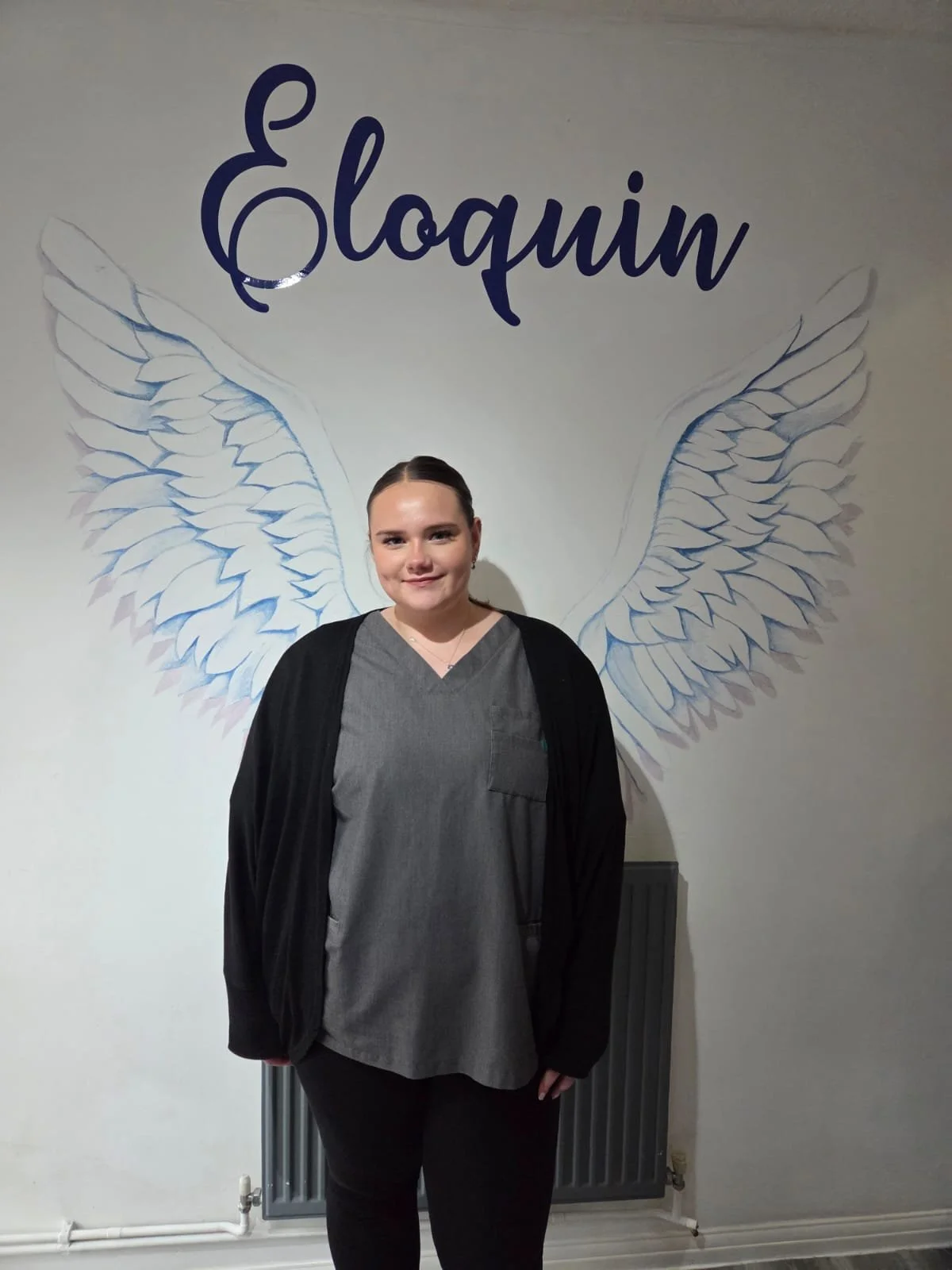 A woman standing in front of a wall with a mural of angel wings and the word 'Elouin' written above.