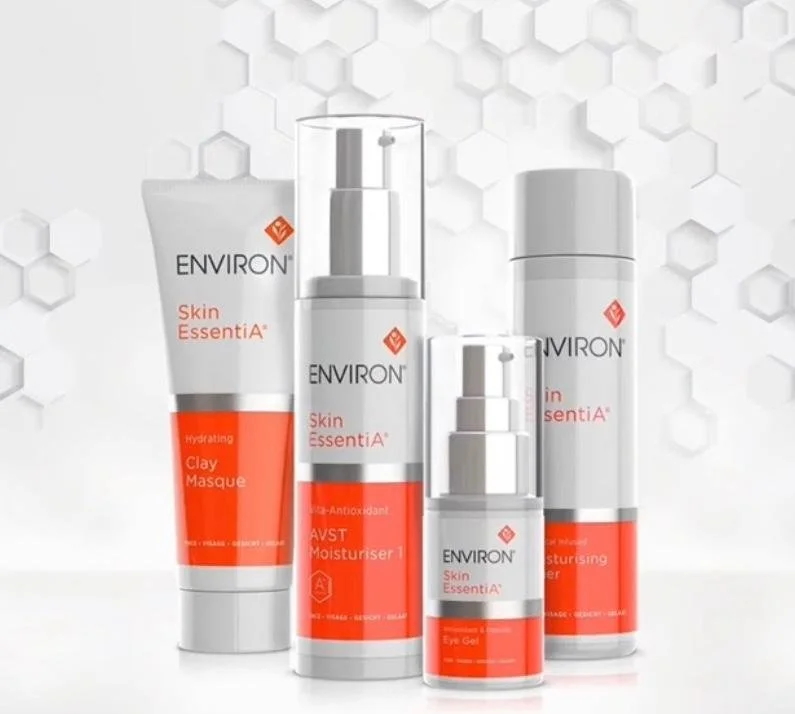 Four bottles and one tube of skincare products from Environ's Skin EssentiA line, including a clay mask, moisturizer, and eye gel, arranged against a white honeycomb pattern background.