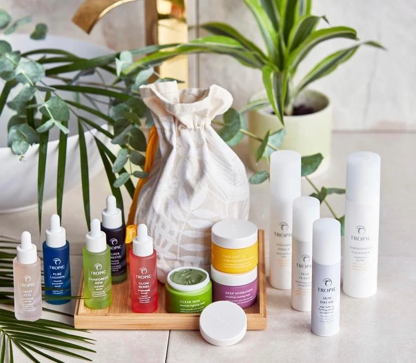 Skincare products and plant on a white table, including bottles, jars, and a cloth bag, with leafy plants in the background.