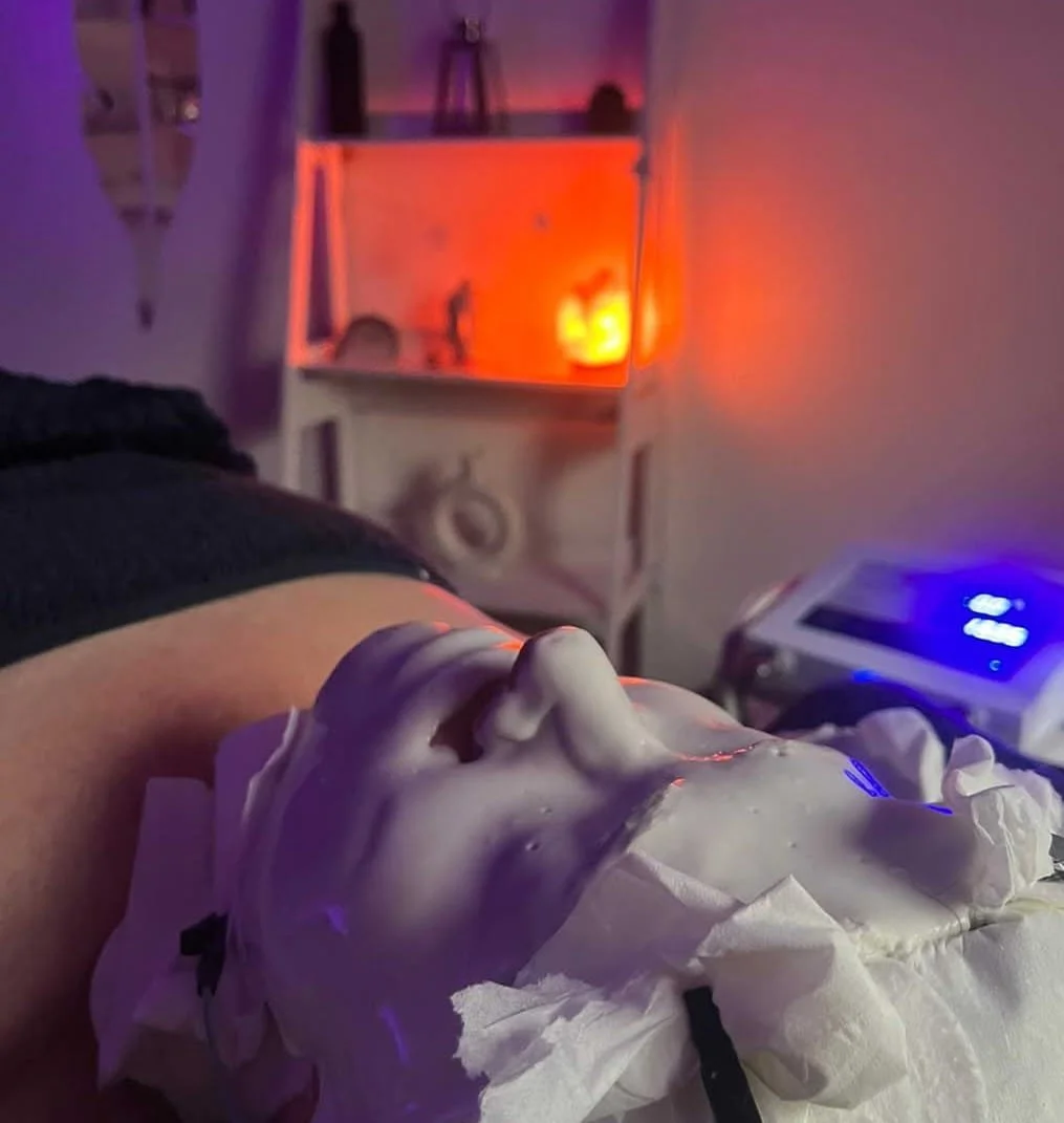 A person receiving a facial treatment with a crazy amount of cream on their face, lying down with a mask on. The background shows a cozy room with purple and orange ambient lighting, shelves, and a machine.