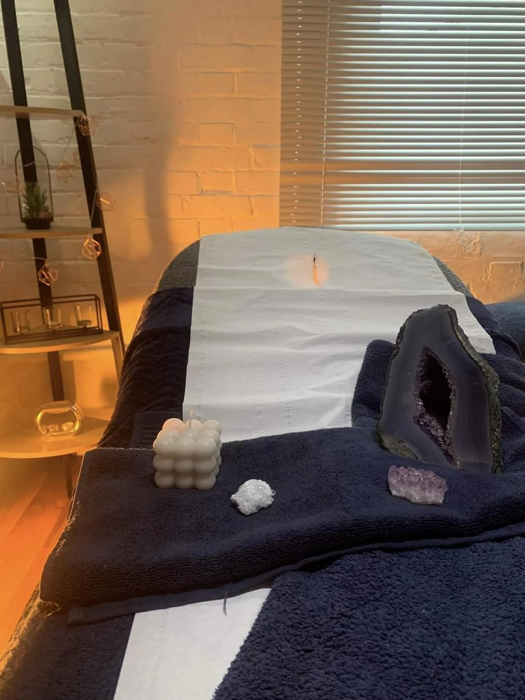 A massage table with a white sheet and decorative stones and crystals on a black towel. The room has a window with blinds, a white brick wall, and a shelving unit with plants and candles.