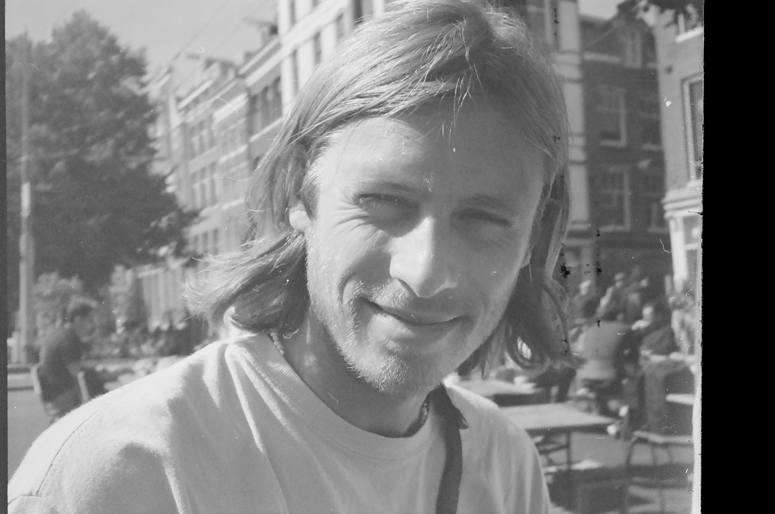 Black and white photo of a smiling man with shoulder-length hair, wearing a T-shirt, outdoors with buildings and trees in the background.