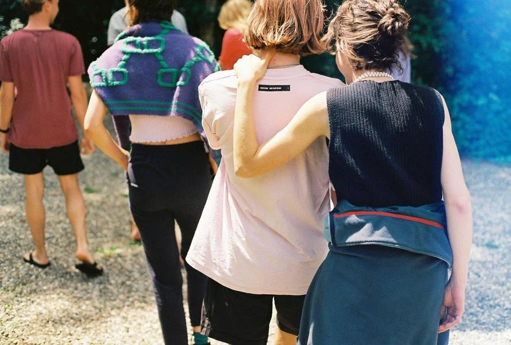 People walking outdoors on a sunny day, with two women arm-in-arm in foreground and others behind them.