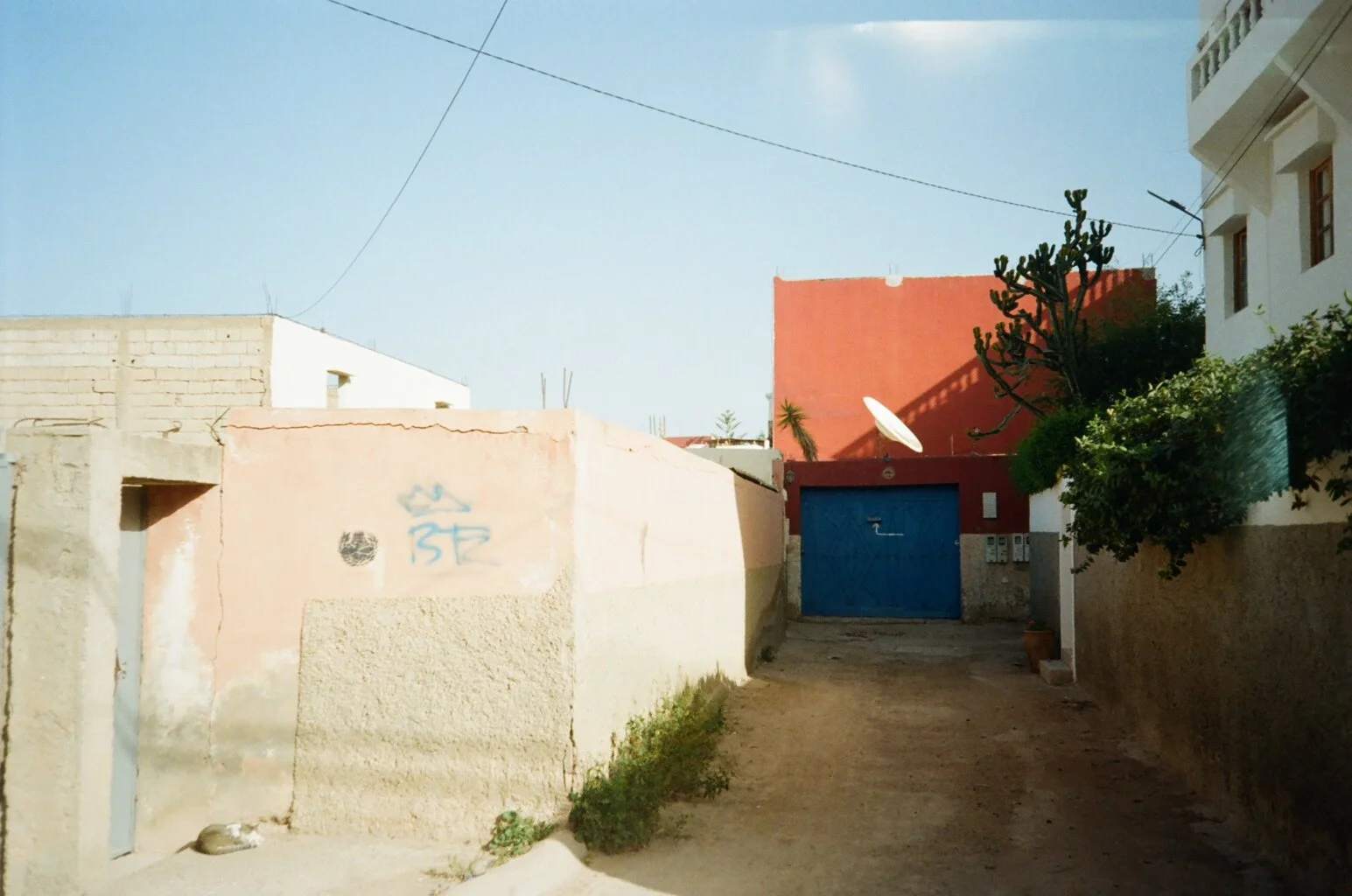 An alleyway in a neighborhood with various buildings, including a pink wall with graffiti, a blue gate, and a red building. There are trees, bushes, and a satellite dish on one building, with clear blue sky overhead.