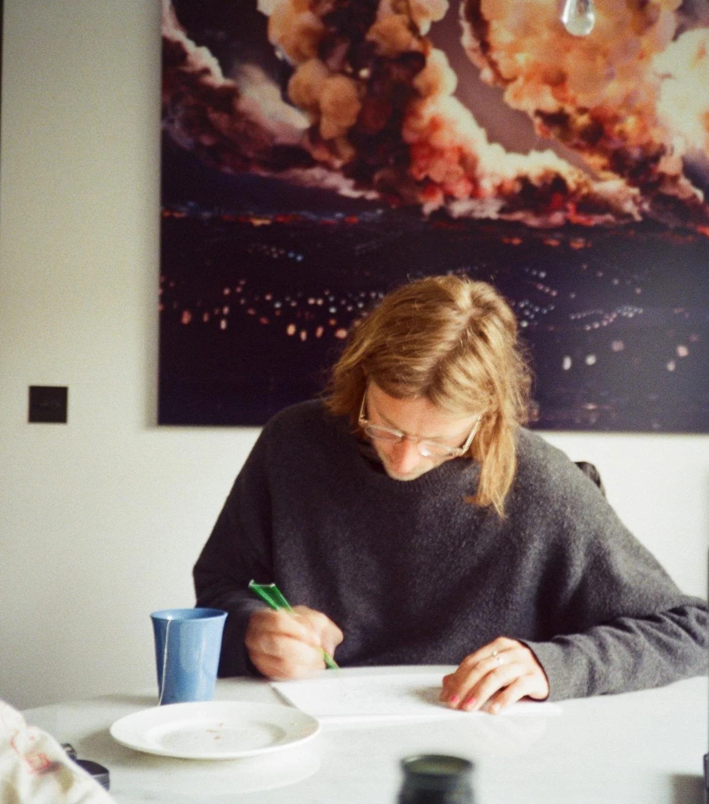 Woman with glasses writing at a table with a white tablecloth, a blue mug, and an empty white plate in front of her, with a large space-themed photograph of clouds and city lights in the background.
