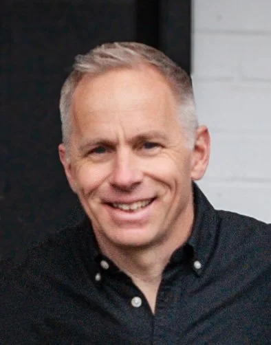 A smiling man with short blond hair wearing a black button-up shirt.