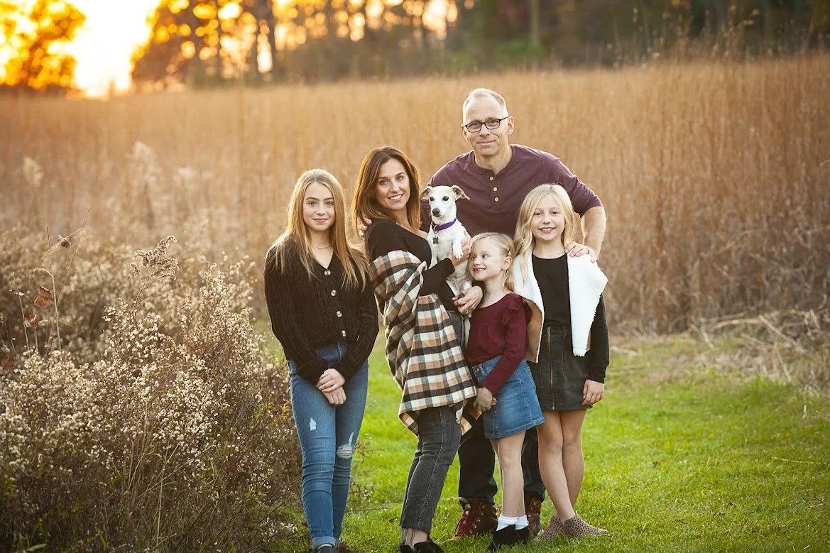 Family of six, including three girls, one woman, one man, and a small dog, standing outdoors in a field during sunset, smiling at the camera.