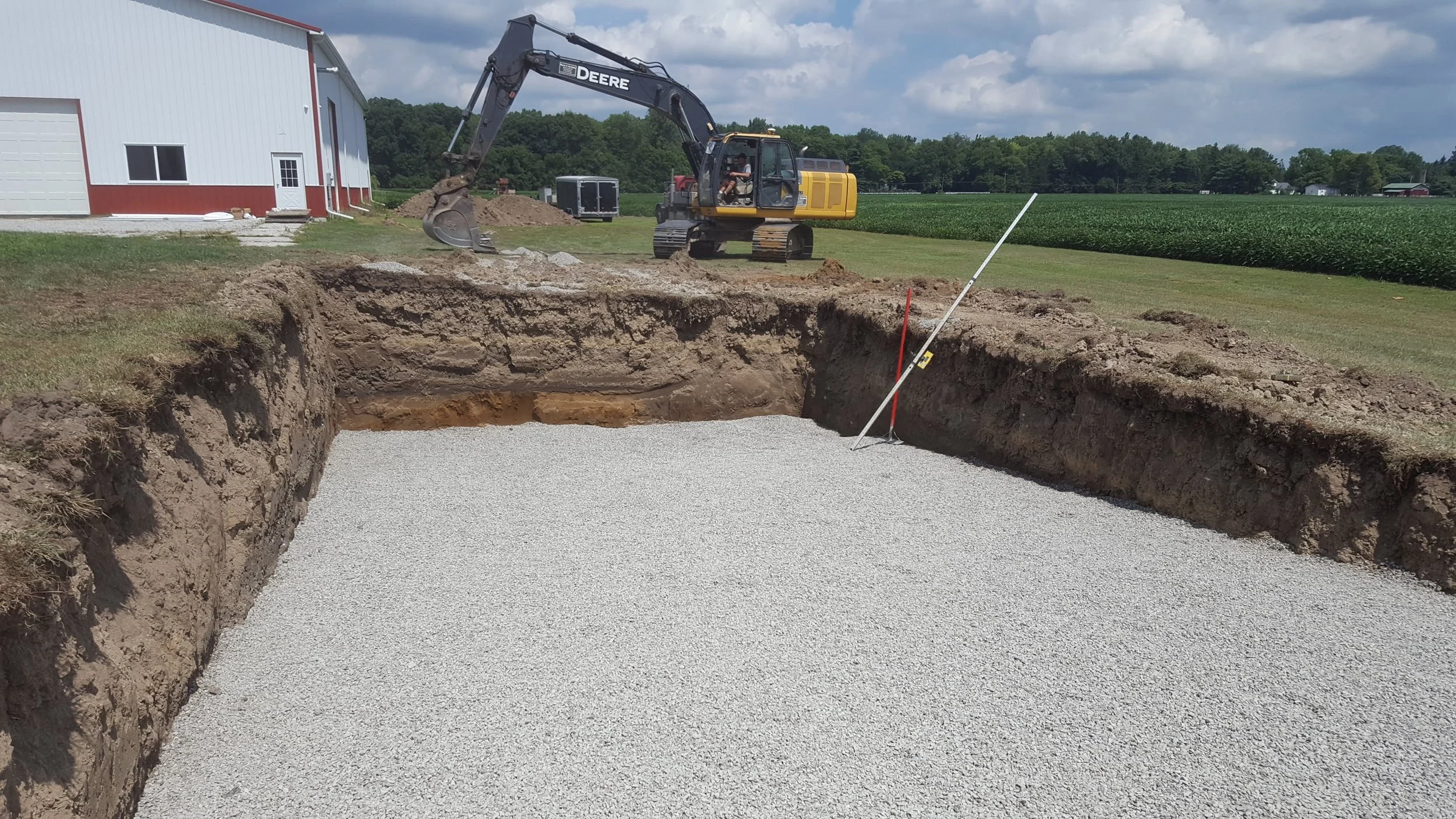 A construction site on a farm with a large excavator digging a hole and a white gravel base inside the hole. A red and white barn is visible in the background along with green fields and trees, under a partly cloudy sky.