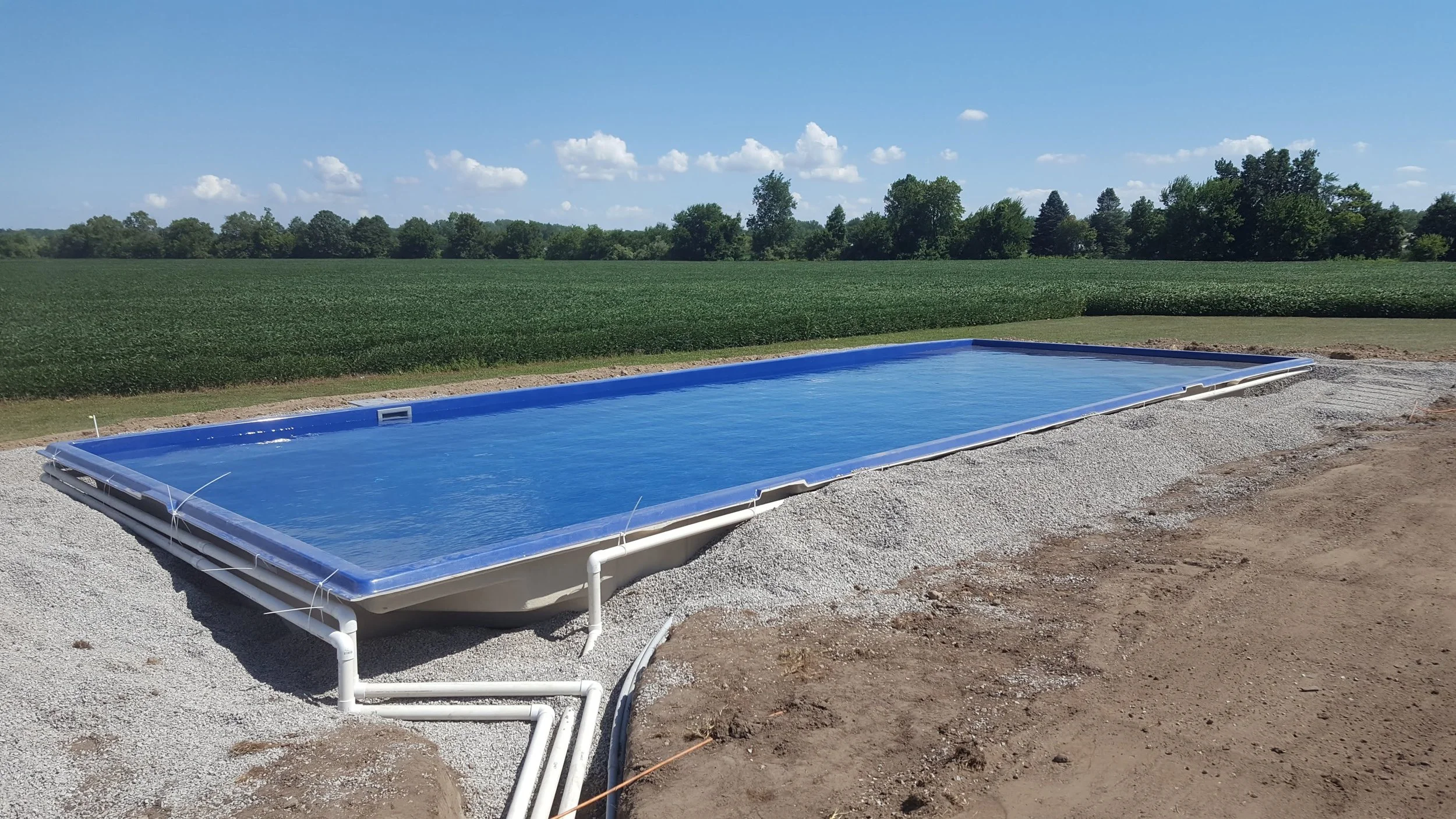 An in-ground rectangular swimming pool filled with water, situated outdoors on a construction site with gravel around it, with a background of green fields and trees under a blue sky with scattered clouds.