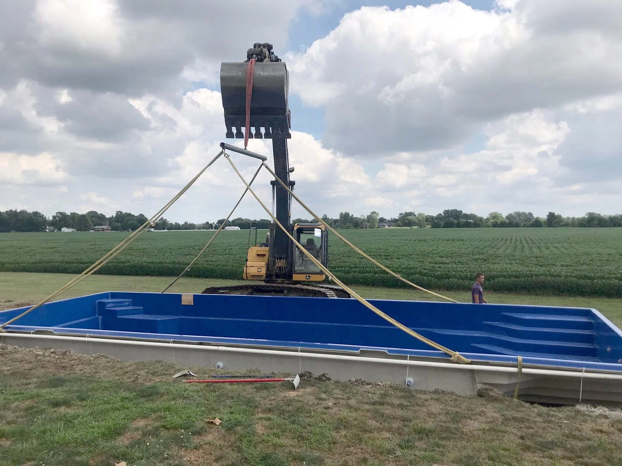 A construction site on a field with a large excavator lifting a metal structure, with safety ropes securing the structure, and a person standing nearby.