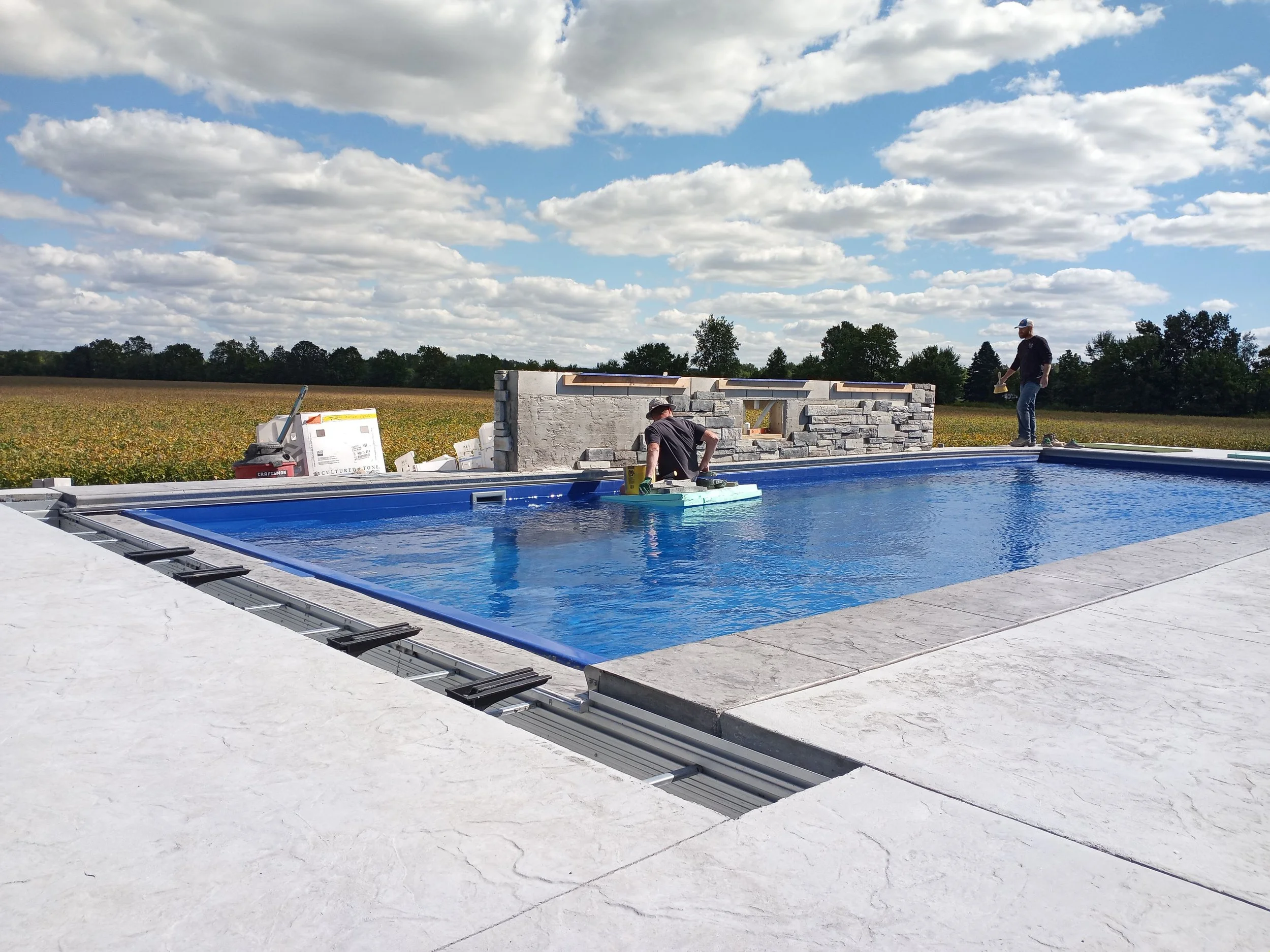 Construction workers building a stone wall near an outdoor swimming pool with a rural landscape and partly cloudy sky in the background.