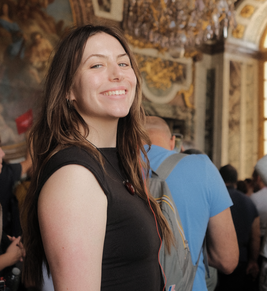 A woman with long brown hair smiling in a crowded indoor space with ornate decorations and a chandelier.