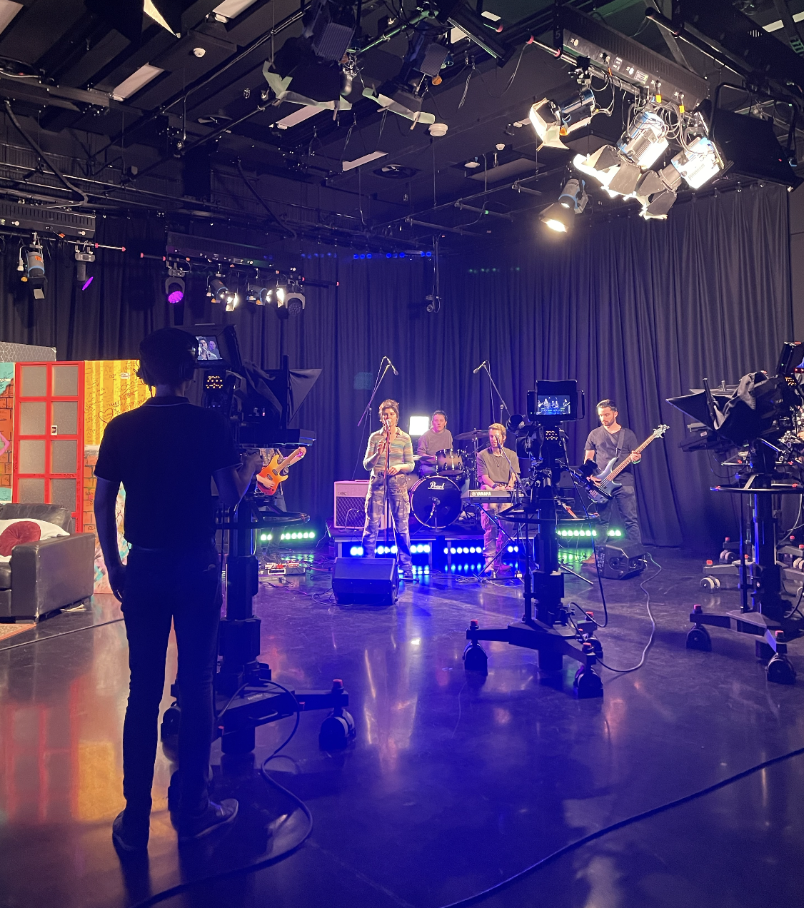 A band rehearsing on a stage with black curtains, with members playing instruments and a singer using a microphone, while a cameraman records them. Stage lighting and professional cameras are also visible.