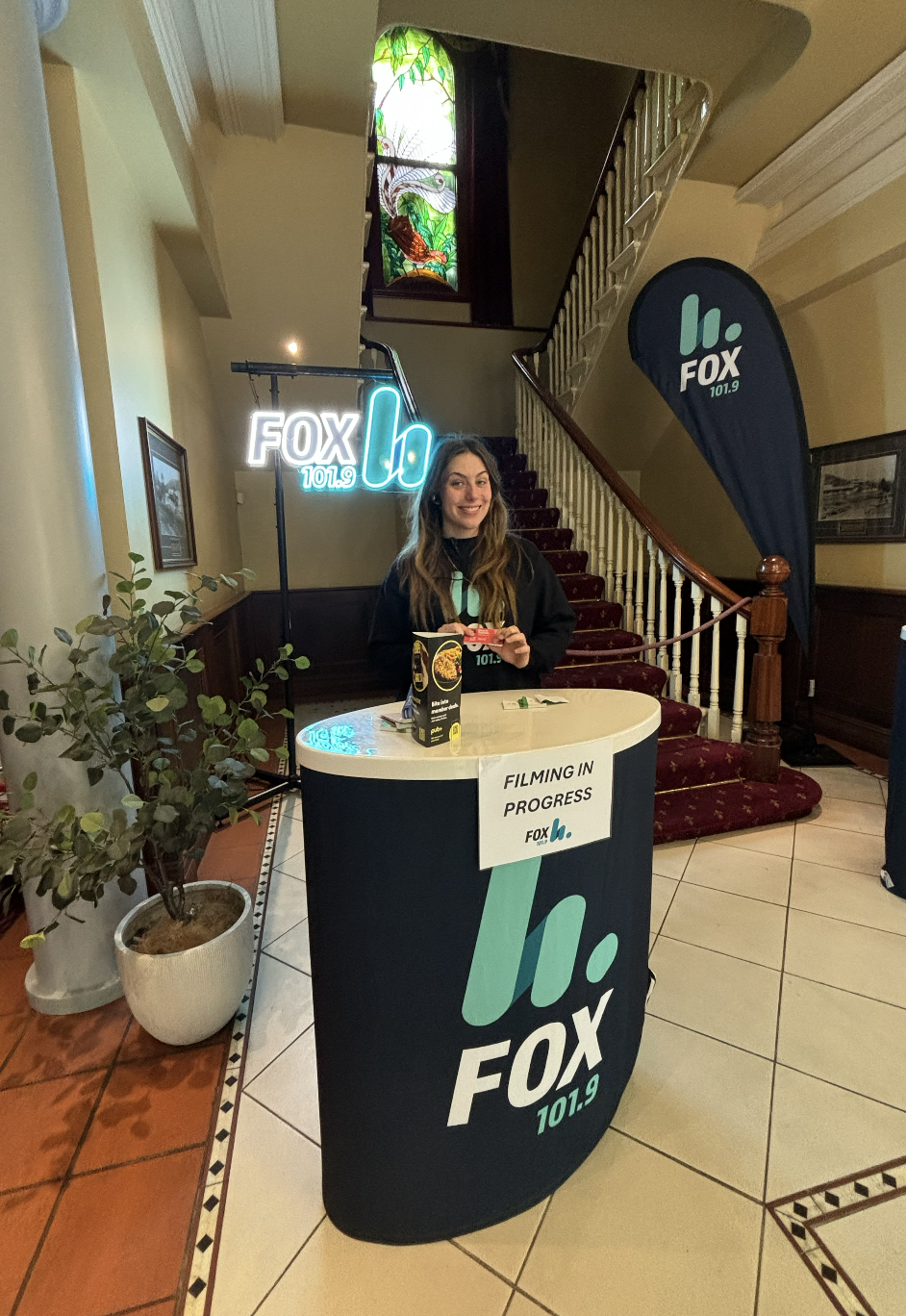 A young woman standing behind a promotional booth for FOX 101.9 radio station inside a building with wooden stairs and stained glass window. The booth has a sign that reads "Filming in Progress" and the woman is smiling, holding a snack. There are banners and signs for FOX 101.9.