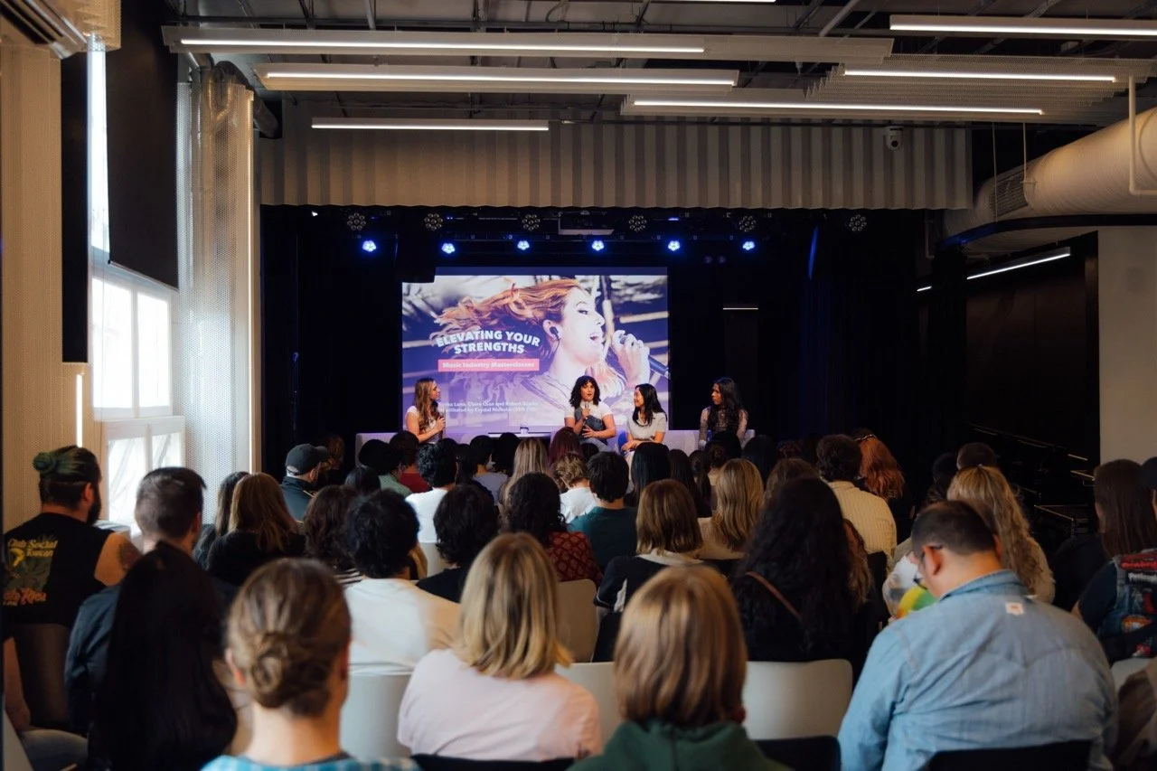 A panel discussion at a conference with four women on stage speaking into microphones, in front of a large screen displaying the text "Elevating Your Strengths" and an image of a woman singing. The audience is seated and listening attentively.