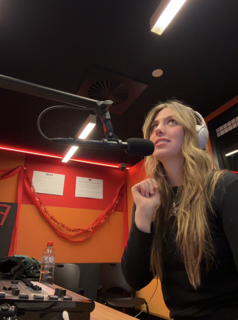 A woman wearing headphones speaking into a microphone in a recording studio with orange and red walls, decorated with tinsel and flags, and a sound mixer on the table.