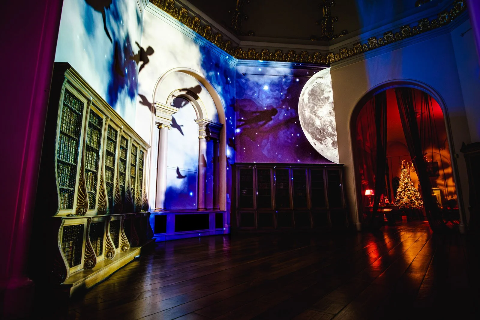A room with a bookshelf on the left, star-shaped shadows on a wall, a large projected image of the moon on the right, and a Christmas tree illuminated with lights in the background through a doorway, creating a festive atmosphere.