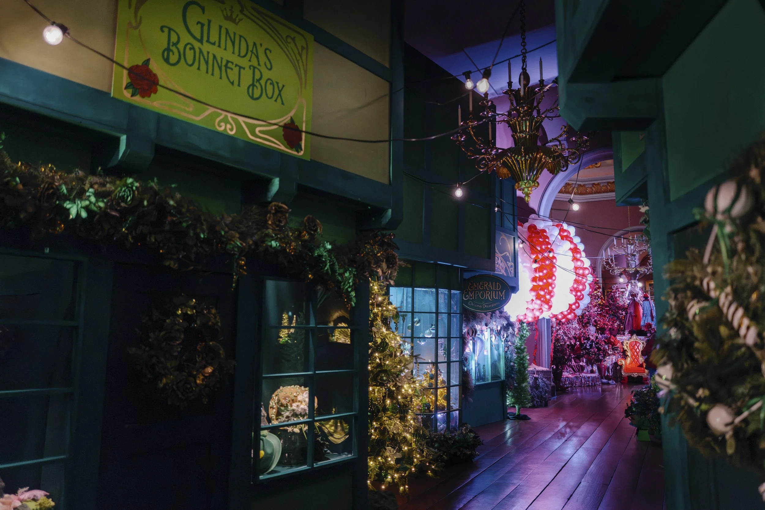 Indoor scene of a vintage shop decorated with Christmas ornaments, lights, and floral displays, with a chandelier hanging from the ceiling and a sign that reads 'Glinda's Bonnet Box'.