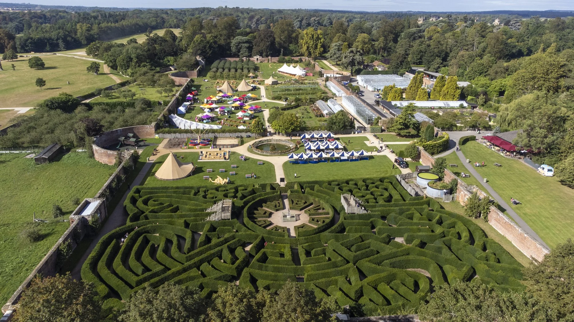 Aerial view of a large outdoor event with tents, a maze garden, greenhouses, and a park surrounded by trees.