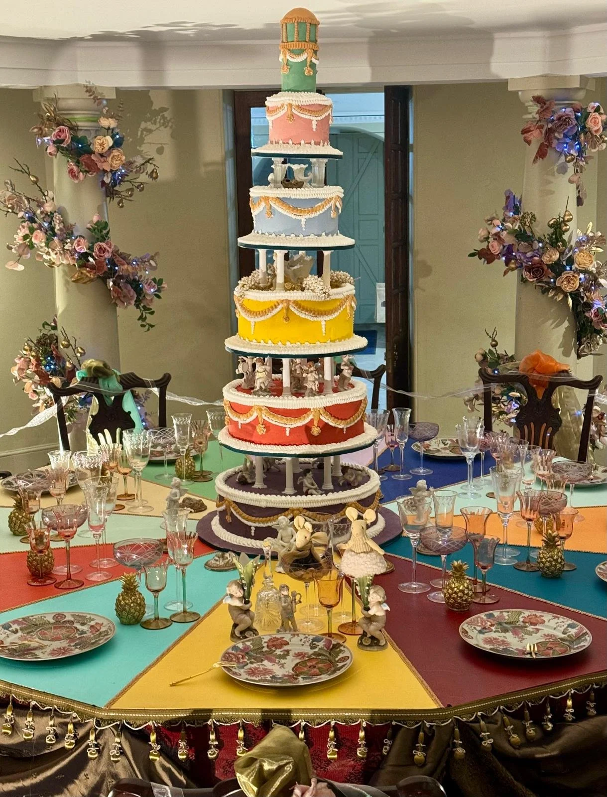 Colorful tiered cake on a table with decorated place settings, surrounded by decorative figurines and pineapple-shaped accents, in a room with floral and ribbon decorations.