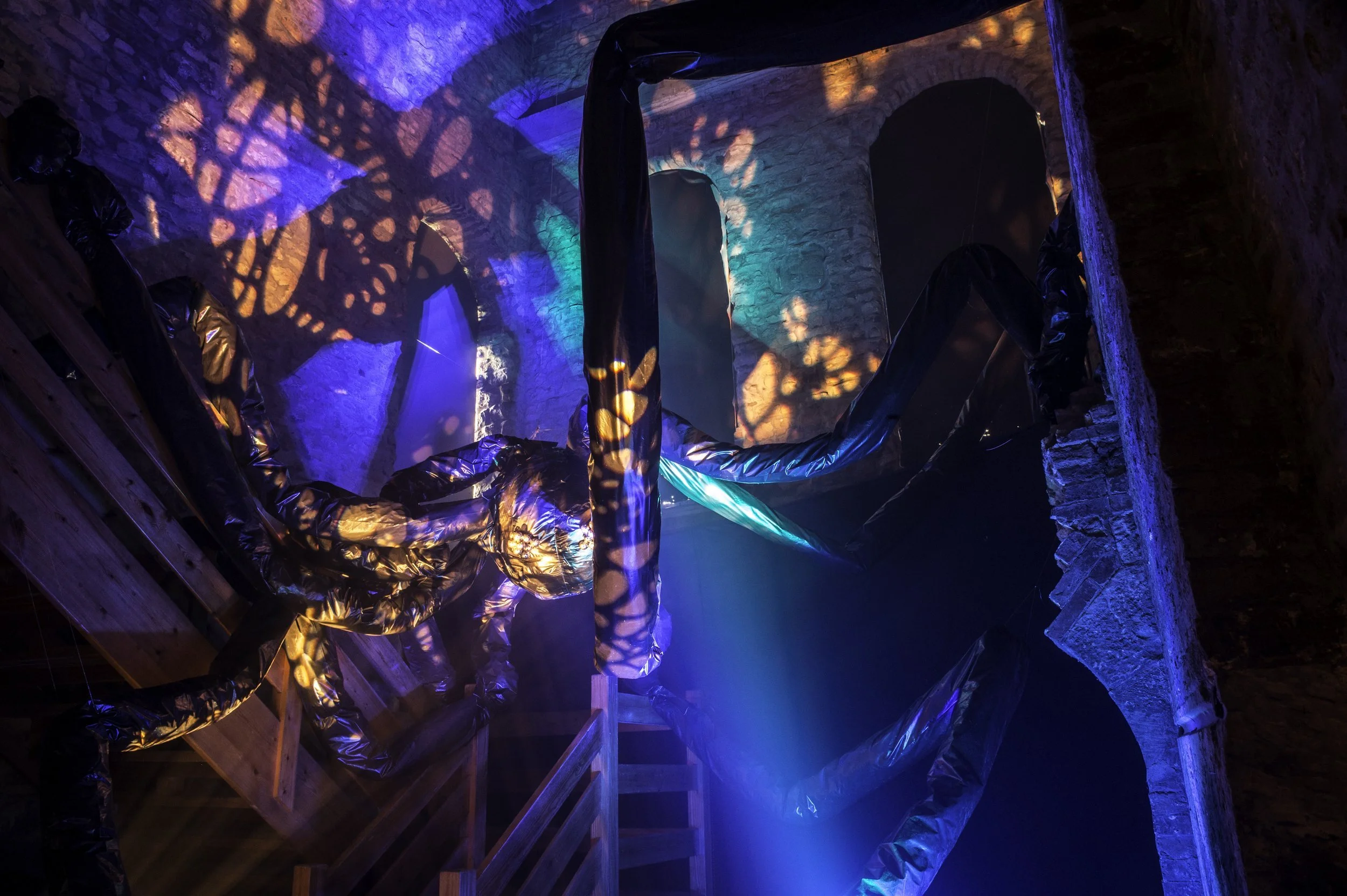 Underground room with brick walls illuminated by colorful lights, with some black plastic and wooden objects on the floor and scaffolding.