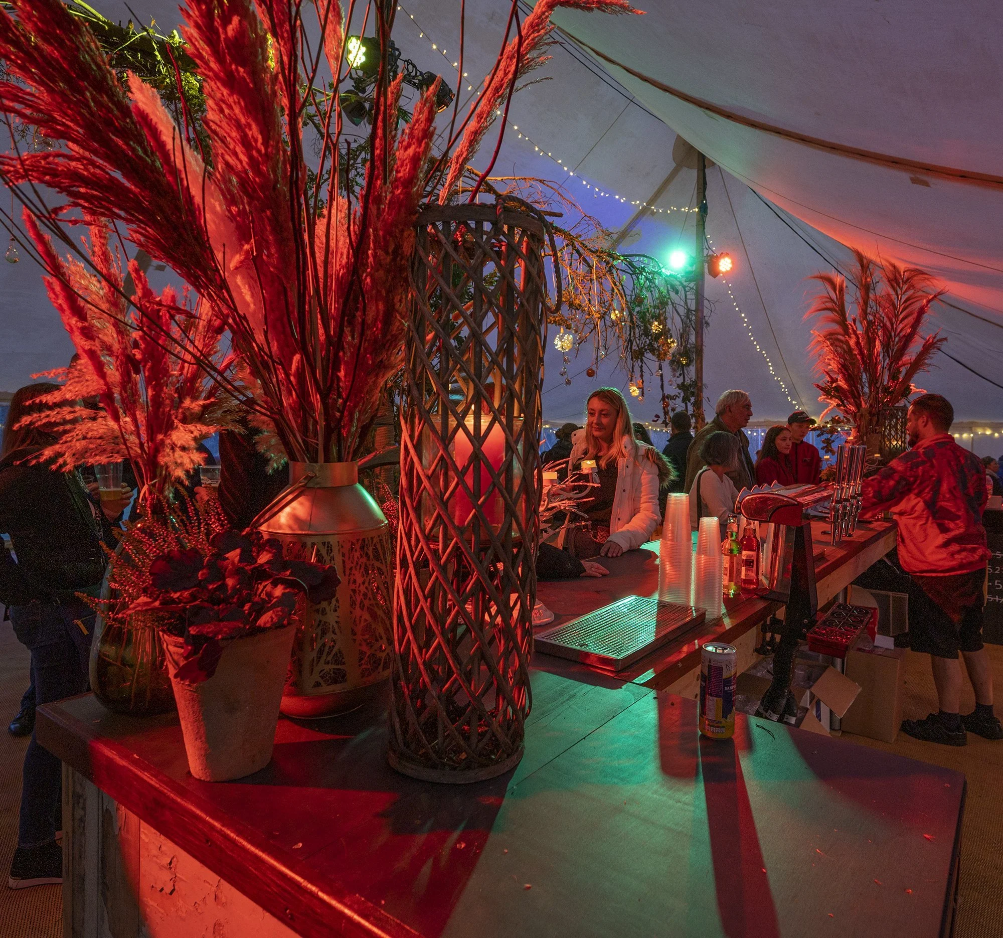 Decorated event tent with red and green lighting, floral arrangements, and people socializing.