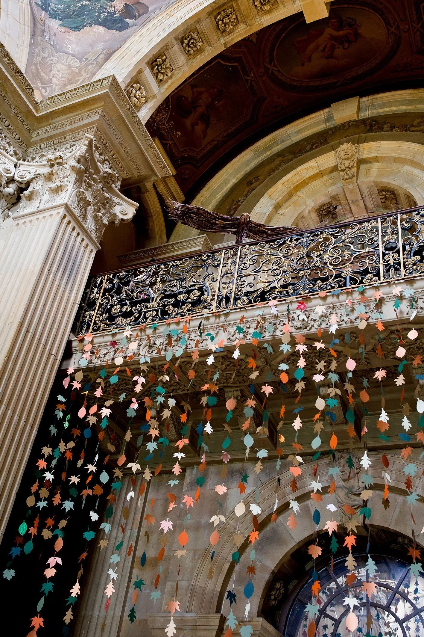 Interior view of a historic building with ornate architectural details, including decorative columns and arches. Hanging paper or fabric leaves in various colors decorate the space, creating a festive atmosphere.