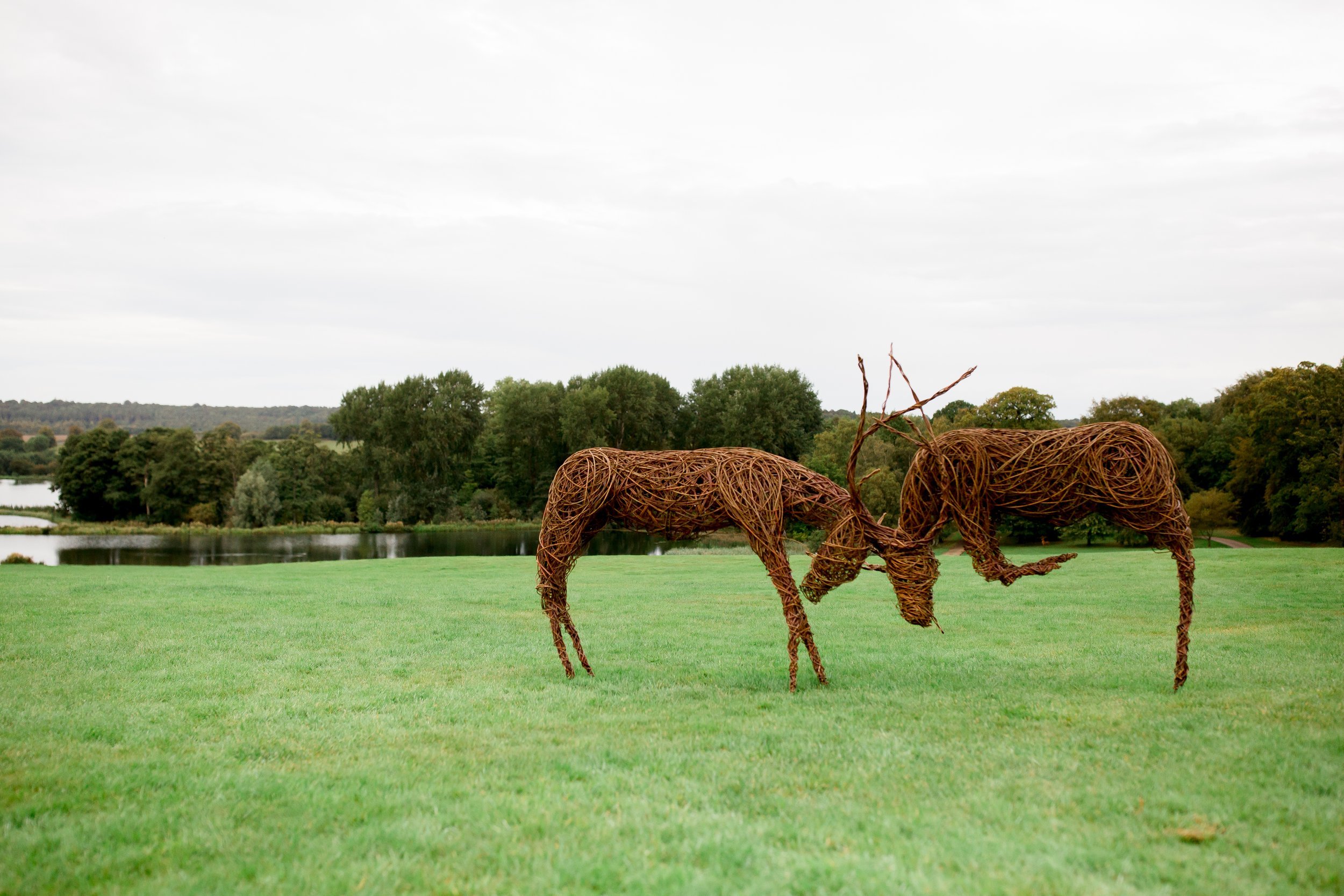 Two animal sculptures made of intertwined twigs on a grassy field with a lake and trees in the background under cloudy sky.
